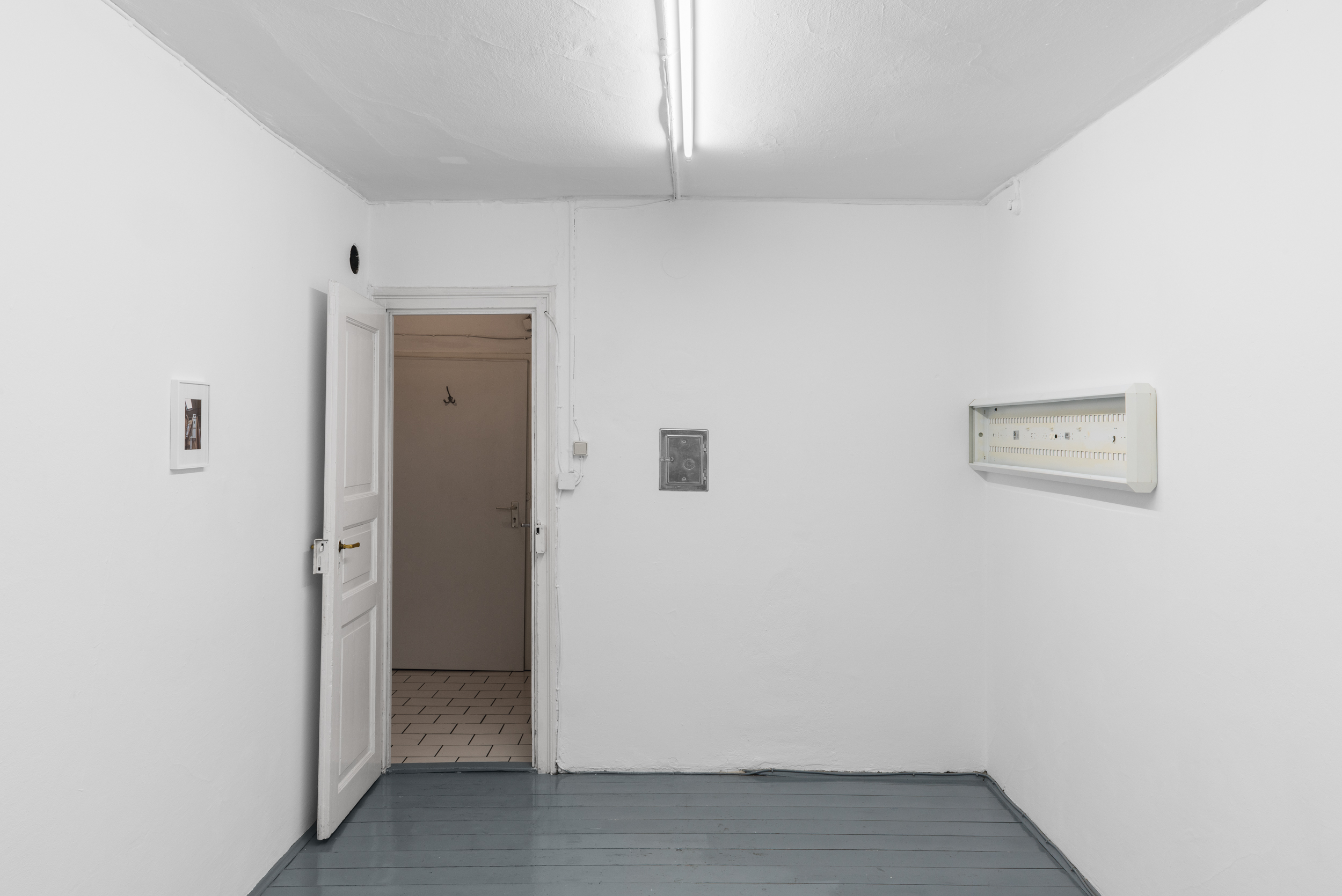 Aisha Altenhofen solo exhibition at Austin Space: Installation view towards the door with a warmer light temperature, a small photographic work hangs on the opposite wall to a grid light fixture that is hung on the wall.